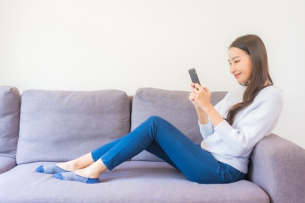 Schöne junge asiatische Frau des Porträts, die intelligentes Handy auf Sofa im Wohnzimmerinnenraum verwendet