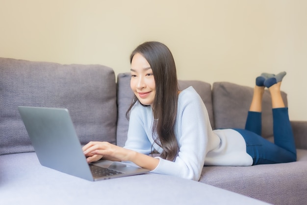 Schöne junge asiatische Frau des Porträts, die Laptop für die Arbeit im Wohnzimmer verwendet