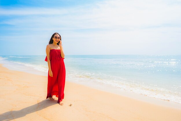 Schöne junge asiatische Frau des Porträts, die Mobiltelefon oder Handy auf dem Strand und dem Meer verwendet