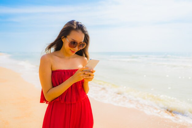 Schöne junge asiatische Frau des Porträts, die Mobiltelefon oder Handy auf dem Strand und dem Meer verwendet