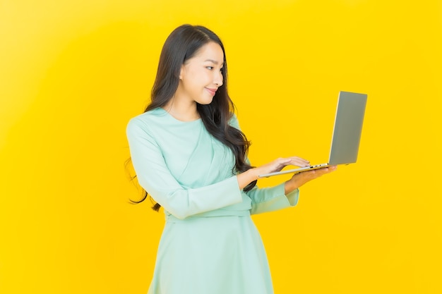 Schöne junge asiatische Frau des Porträts lächeln mit Computerlaptop auf Gelb