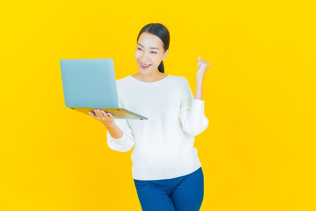 Schöne junge asiatische Frau des Porträts lächeln mit Computerlaptop auf Gelb