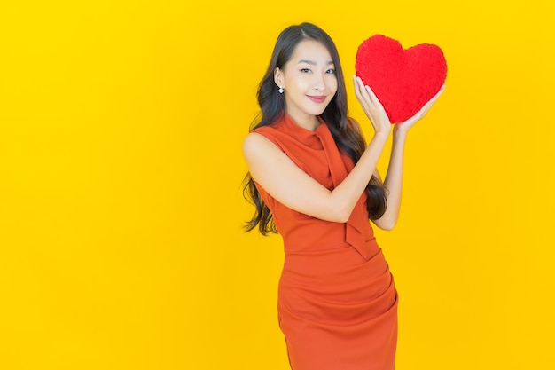 Schöne junge asiatische Frau des Porträts lächeln mit Herzkissenform auf Gelb