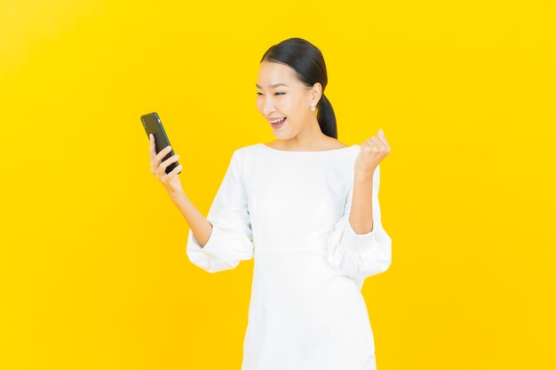 Schöne junge asiatische Frau des Porträts lächeln mit intelligentem Handy auf Gelb