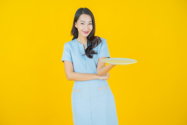 Schöne junge asiatische Frau des Porträts lächeln mit leerem Tellerteller auf Farbhintergrund