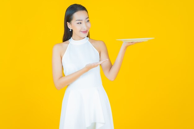Schöne junge asiatische Frau des Porträts lächeln mit leerem Tellerteller auf Gelb