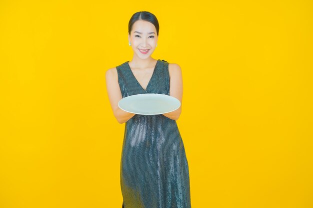 Schöne junge asiatische Frau des Porträts lächeln mit leerem Tellerteller auf Gelb