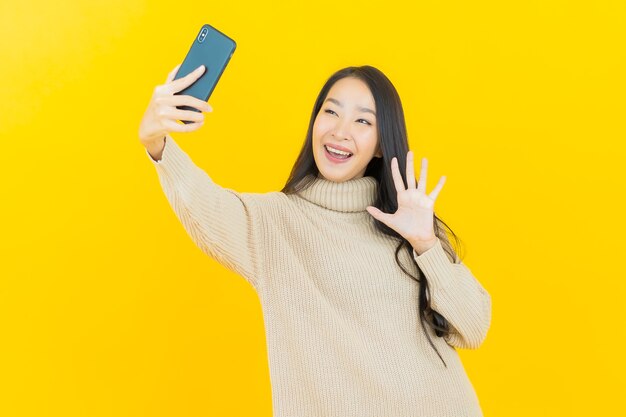 Schöne junge asiatische Frau des Porträts lächelt mit intelligentem Handy auf gelber Wand