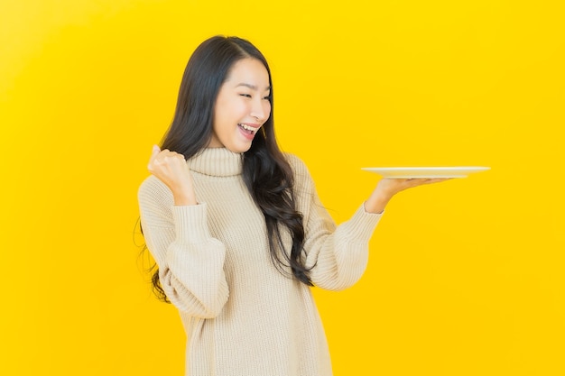 Schöne junge asiatische Frau des Porträts lächelt mit leerem Tellerteller auf gelber Wand