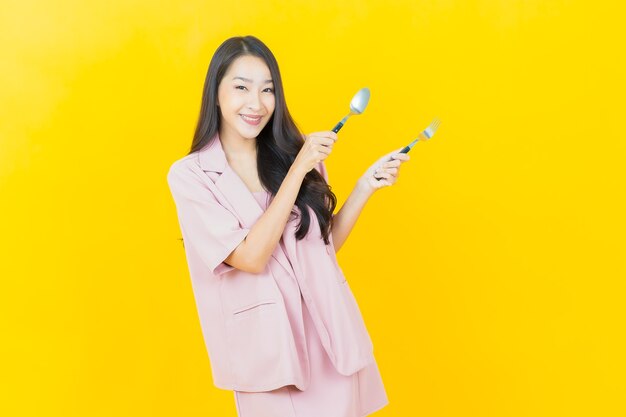 Schöne junge asiatische Frau des Porträts lächelt mit Löffel und Gabel auf gelber Wand