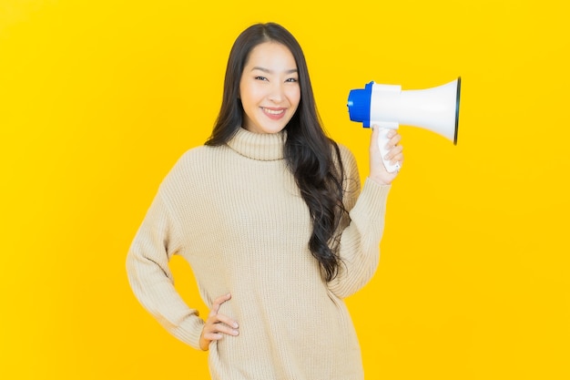 Schöne junge asiatische Frau des Porträts lächelt mit Megaphon auf gelber Wand