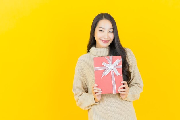 Schöne junge asiatische Frau des Porträts lächelt mit roter Geschenkbox auf gelber Wand