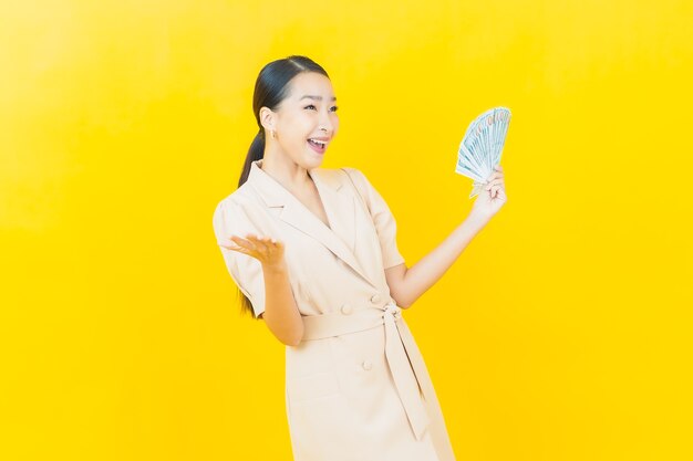 Schöne junge asiatische Frau des Porträts lächelt mit viel Bargeld und Geld auf Farbwand