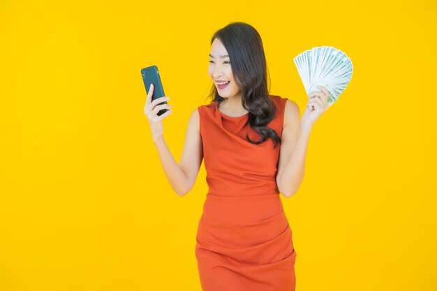 Schöne junge asiatische Frau des Porträts lächelt mit viel Bargeld und Geld auf Gelb