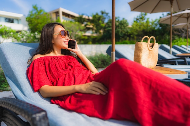 Schöne junge asiatische Frau des Porträts mit Handy oder Handy um Swimmingpool am Hotelerholungsort
