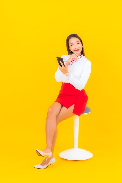 Schöne junge asiatische Frau des Porträts mit intelligentem Handy auf Gelb