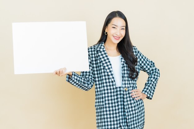 Schöne junge asiatische Frau des Porträts mit leerer weißer Anschlagtafel auf Beige