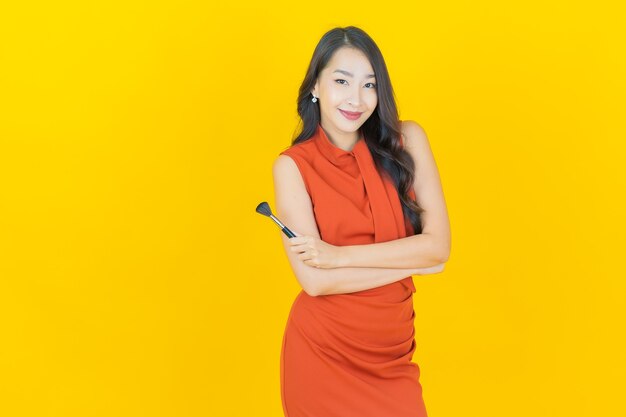 Schöne junge asiatische Frau des Porträts mit Make-upbürstenkosmetik auf Gelb