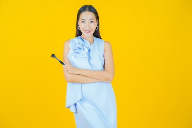 Schöne junge asiatische Frau des Porträts mit Make-upbürstenkosmetik auf Gelb