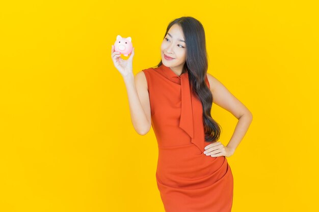 Schöne junge asiatische Frau des Porträts mit Sparschwein auf Gelb
