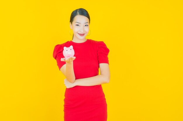 Schöne junge asiatische Frau des Porträts mit Sparschwein auf Gelb