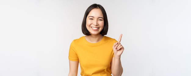 Schöne junge asiatische Frau, die mit dem Finger nach oben zeigt, lächelt und amüsiert in die Kamera schaut, die Werbung zeigt ...