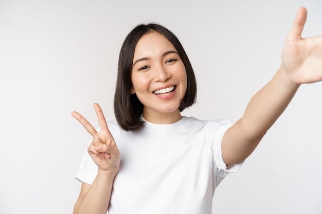 Schöne junge asiatische frau, die selfie macht, das mit dem friedenszeichen posiert, das glücklich lächelt, macht ein foto, das vor weißem hintergrund aufwirft