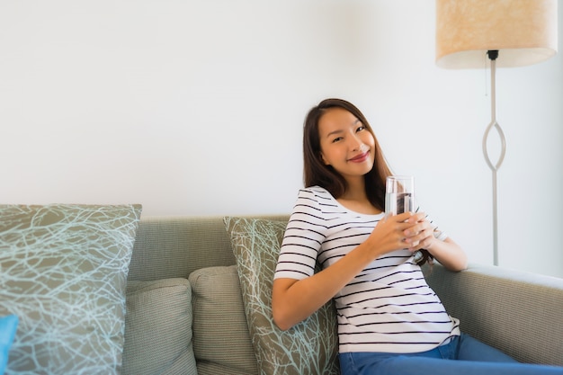 Schöne junge asiatische Frauen des Porträts, die Getränkwasserglas auf Sofa halten