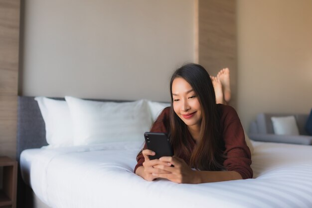 Schöne junge asiatische Frauen des Porträts, die Handy auf Bett verwenden