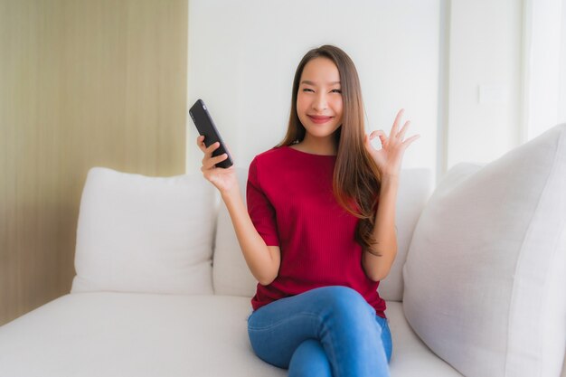 Schöne junge asiatische Frauen des Porträts, die Mobile oder intelligentes Telefon auf Sofa verwenden