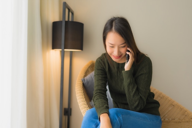 Schöne junge asiatische Frauen des Porträts, die Unterhaltungshandy verwenden und auf Sofastuhl sitzen