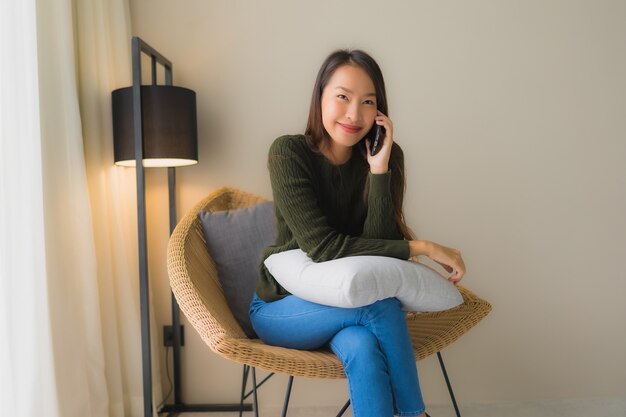 Schöne junge asiatische Frauen des Porträts, die Unterhaltungshandy verwenden und auf Sofastuhl sitzen