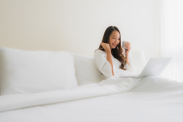 Schöne junge asiatische Frauen des Porträts mit Kaffeetasse- und Computerlaptop auf Bett