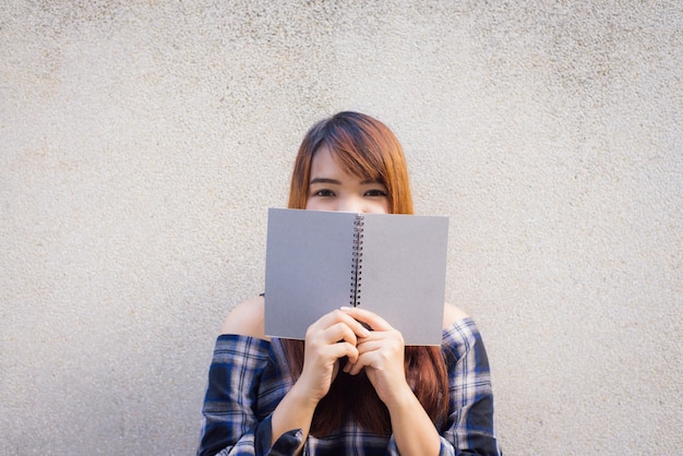 Schöne junge asiatische Frauen versteckt sich hinter einem grauen Buch auf Betonwand Hintergrund. Vintage Effekt Stil Bilder.