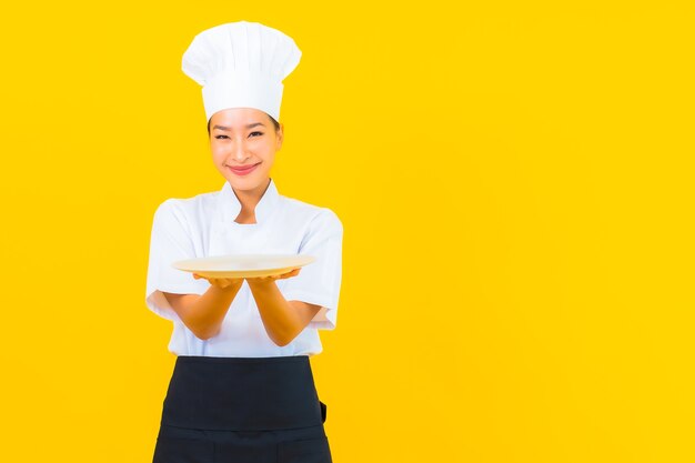 Schöne junge asiatische Kochfrau des Porträts mit Teller auf gelbem lokalisiertem Hintergrund