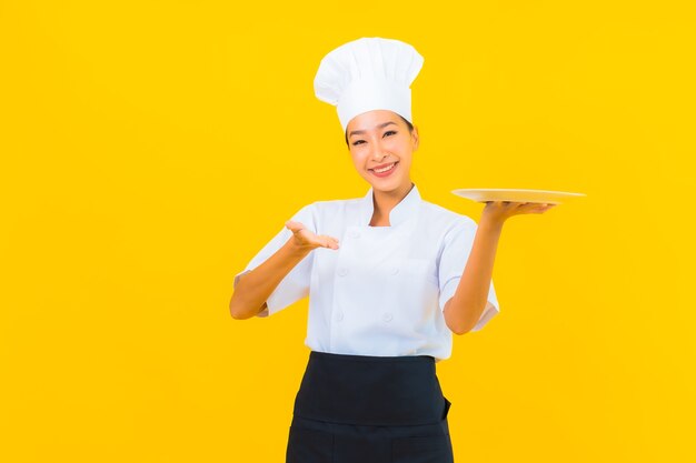 Schöne junge asiatische Kochfrau des Porträts mit Teller auf gelbem lokalisiertem Hintergrund