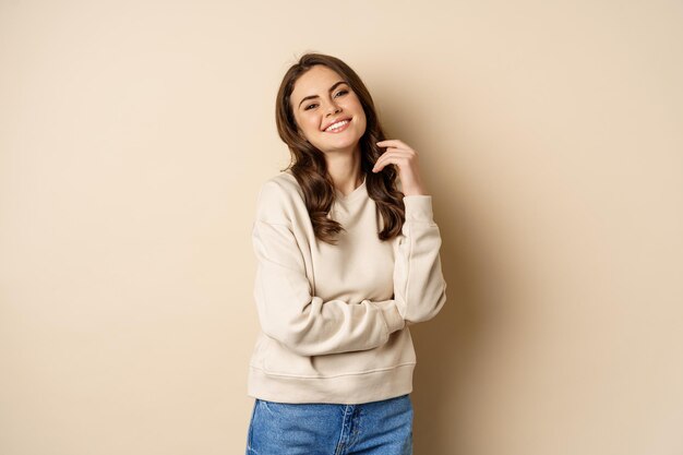 Schöne junge brünette Frau posiert glücklich vor beigem Hintergrund und lächelt in die Kamera mit Pullover