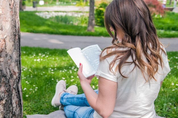 Schöne junge Brünette sitzt auf einem frischen Frühling in einem Park, ein Buch zu lesen. Nahaufnahme einer schönen jungen Frau liest Buch im Park. Frau, die ein Buch liest, das draußen liest
