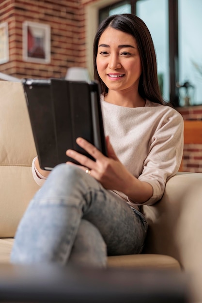 Schöne junge Chinesin, die zu Hause ein digitales Tablet verwendet, das Internet Web 3.0 lächelnd anschließt, während sie auf einem Couch-Unterhaltungsgerät sitzt und mit dem Computer-Tablet eines drahtlosen Geräts im Internet surft