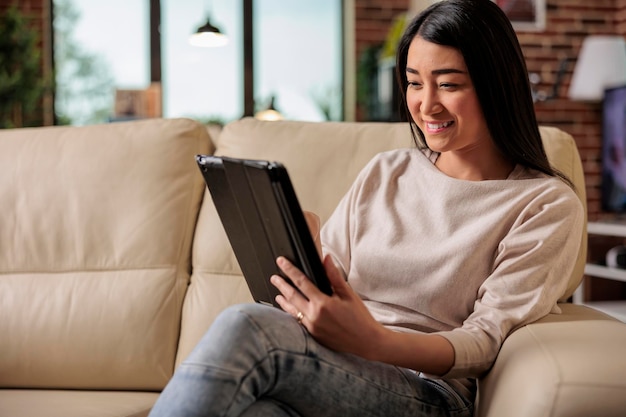 Schöne junge Chinesin, die zu Hause ein digitales Tablet verwendet, das Internet Web 3.0 lächelnd anschließt, während sie auf einem Couch-Unterhaltungsgerät sitzt und mit dem Computer-Tablet eines drahtlosen Geräts im Internet surft