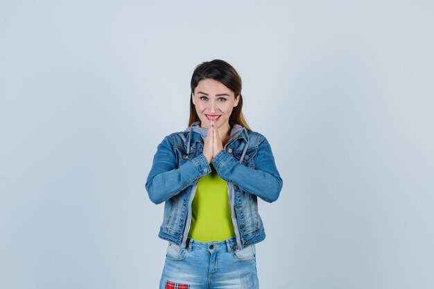 Schöne junge Dame, die gefaltete Hände in flehender Geste in Denim-Outfit zeigt und fröhlich aussieht. Vorderansicht.