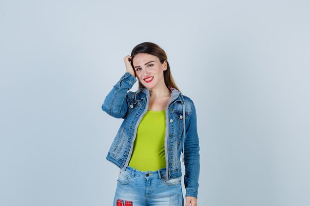 Schöne junge Dame, die im Denim-Outfit die Hand auf dem Kopf hält und jovial aussieht, Vorderansicht.