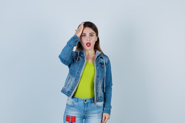 Schöne junge Dame in Denim-Outfit, die die Hand auf dem Kopf hält und vergesslich aussieht, Vorderansicht.