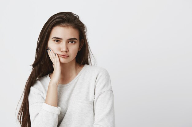 Schöne junge dunkeläugige Frau mit dunklem langem Haar, das losen Pullover trägt, Hand auf Wange hält und mit ernstem Ausdruck schaut