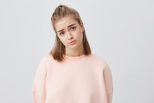 Schöne junge europäische Frau mit hellem Haar, das mit unglücklichem und bedauerndem Blick schaut. Trauriges hübsches Mädchen, das sich verärgert fühlt, während es Zeit allein zu Hause verbringt.