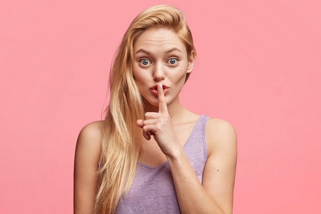 Schöne junge Frau blonde junge Frau hält Vorderfinger auf den Lippen, erzählt vertrauliche Informationen, fordert völlige Stille