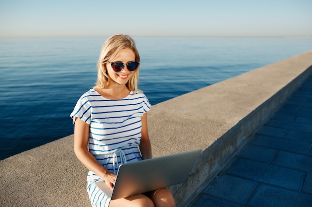 Schöne junge Frau, die am Strand mit Laptop sitzt