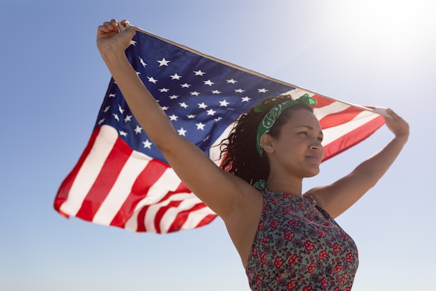 Schöne junge Frau, die amerikanische Flagge auf Strand im Sonnenschein wellenartig bewegt