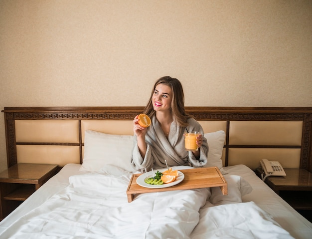 Schöne junge Frau, die auf dem Bett isst Obst und Saft im Frühstück sitzt