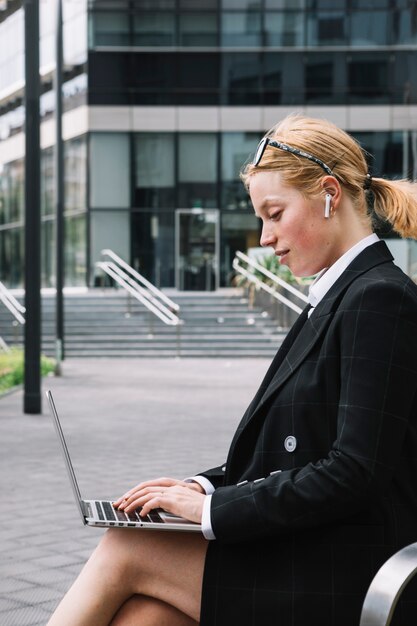 Schöne junge Frau, die außerhalb des Bürogebäudes schreibt auf Laptop sitzt